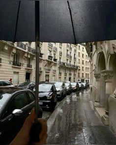 a person holding an umbrella in the rain near parked cars on a city street with tall buildings