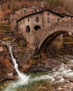 an old stone bridge over a river with a waterfall running under it and buildings on the other side