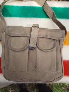 "This simple military style waxed canvas shoulder bag is made from heavy 100% cotton canvas and is treated with a beeswax blend that repels water; treating canvas with wax is a method used since the 1920's to make the material water resistant. It features one large main compartment and two front pockets with hook and loop closures. The main flap closes with a strap and a metal fold over style buckle. The bag measures approximately 10\" across by 8\" tall and 3.5\" wide with a strong 1.5\" wide a Khaki Waxed Canvas Shoulder Bag For Outdoor, Khaki Canvas Shoulder Bag With Canvas Lining, Khaki Shoulder Canvas Bag For Outdoor Activities, Khaki Waxed Canvas Shoulder Bag With Adjustable Strap, Outdoor Khaki Waxed Canvas Shoulder Bag, Khaki Canvas Satchel, Outdoor Khaki Canvas Satchel, Canvas Utility Shoulder Bag For Outdoor, Utility Canvas Shoulder Bag For Outdoor