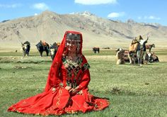 a woman dressed in red sitting on the grass with cows behind her and mountains in the background