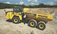 a large yellow dump truck parked on top of a dirt covered field with trees in the background