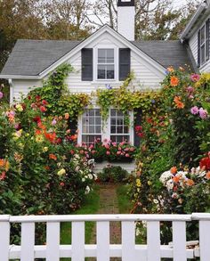 a white house surrounded by flowers and greenery with a white picket fence in front