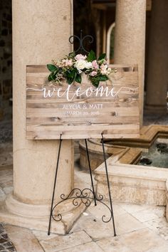 a wooden sign sitting on the side of a building next to a water fountain with flowers growing out of it