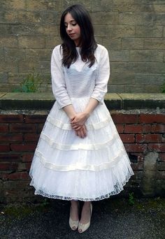 a woman standing in front of a brick wall wearing a white dress with tiered layers