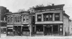 an old black and white photo of two buildings on the corner of a street in front of some shops