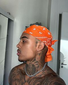 a man with tattoos and an orange bandana stares into the distance while standing in front of a mirror