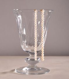 Two long earrings that are strands of pinkish pearls hang off a crystal glass in front of a grey background. The 8, A Line, Angeles, Yellow Gold, Gold, Los Angeles