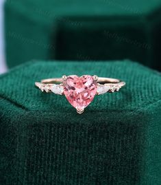 a pink heart shaped diamond ring with three diamonds on the band and an emerald green velvet box