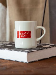 a white coffee cup sitting on top of a table next to a book with a red label