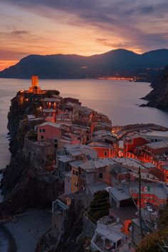 the city is lit up at night by the water's edge with mountains in the background