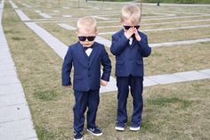 two young boys in suits and sunglasses stand on the grass with their hands to their ears