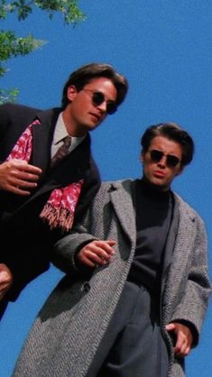 two men standing next to each other in front of a tree and blue sky with one man wearing sunglasses