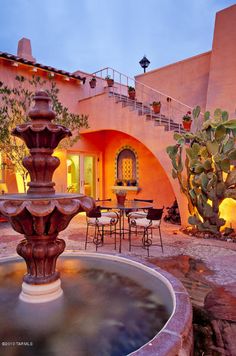 a fountain in the middle of a courtyard with tables and chairs around it at dusk