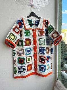 a crocheted jacket hanging on a wall next to a window with a tree in the background