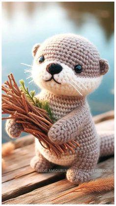 a crocheted stuffed otter holding a twig and looking at the camera while sitting on a dock