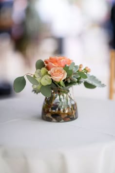 an arrangement of flowers and greenery in a vase on a table with white cloth