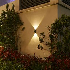 a wall light that is on the side of a building with bushes and trees around it