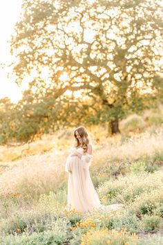 a pregnant woman in a white dress standing in a field with flowers and trees behind her