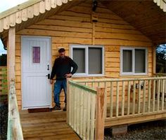 a man standing on the porch of a small wooden cabin with white doors and windows