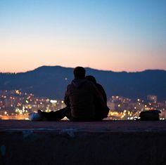 two people sitting on a ledge looking at the city lights in the distance with their backs to each other