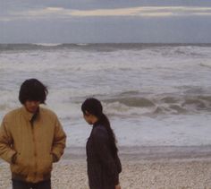 two people standing on the beach next to the ocean looking at something in the distance