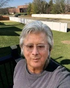 an older woman with glasses sitting on a bench in the grass and looking at the camera