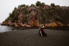 a man and woman standing on top of a sandy beach next to the ocean holding each other