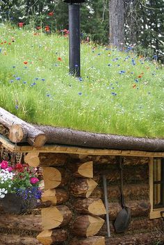 a green roof on top of a log house with flowers in the foreground and trees in the background