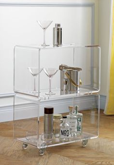 a clear bar cart with bottles and glasses on the bottom, sitting in front of a white wall