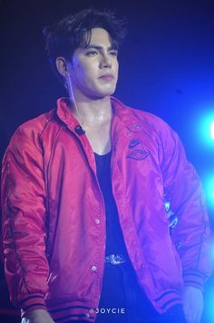 a male in a red jacket and black shirt is standing on stage with his hands in his pockets