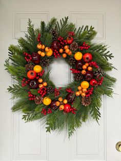 a christmas wreath with fruit and pine cones hanging on a white front door decorated with greenery