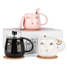 two coffee mugs with faces on them sitting in front of a box and one has a cat's face painted on it