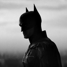 the dark knight batman is standing with his head turned to the side, in front of an overcast sky