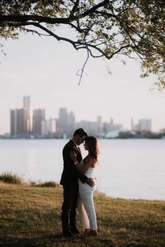 a man and woman standing next to each other under a tree in front of a body of water