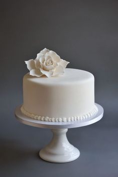 a white cake with a flower on the top is sitting on a stand against a gray background