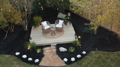 a patio with chairs and lights in the middle of it is surrounded by trees, shrubs, and grass