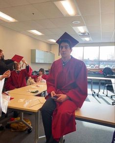 a man in a red graduation gown sitting on a table with other people behind him