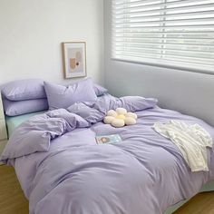 an unmade bed with purple sheets and pillows on top of it next to a window