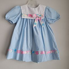 "This adorable vintage dress by Rare Editions is perfect for spring! The dress is made of 100% cotton blue chambray denim. There are short sleeves and buttons at back of neck. There is a square white eyelet collar with floral grosgrain ribbon trim and bow. The trim is mimicked at the hem. Size is: 3t Length: 19.5\" chest: 14\"" Retro Spring Playtime Dresses, Spring Blue Vintage Dress, Blue Retro Cotton Dresses, Retro Blue Cotton Dresses, Light Blue Vintage Cotton Dress, Vintage Light Blue Cotton Dress, Blue Cotton Vintage Dress For Summer, Spring Blue Cotton Vintage Dress, Blue Cotton Vintage Summer Dress