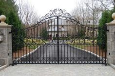 an iron gate in front of a driveway with trees and grass on both sides, surrounded by stone pillars