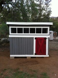 a small gray and red shed sitting in the middle of a dirt field next to trees