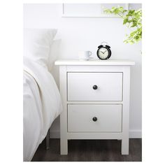 a white nightstand sitting next to a bed with a plant on it and an alarm clock