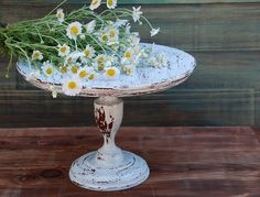 daisies and other flowers are placed on an old cake plate with wood planks in the background