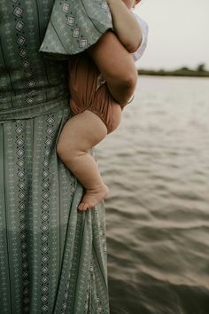 a woman holding a baby in her arms near the water's edge, while wearing a dress