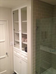 a walk in shower sitting next to a white cabinet with glass doors and shelves filled with towels