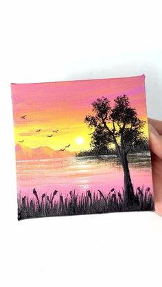 a hand holding up a small canvas with a sunset on the water and birds flying in the sky