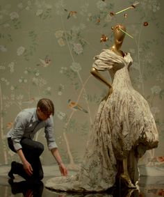 a man kneeling down next to a mannequin in front of a floral wallpaper