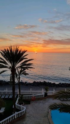 the sun is setting over the ocean with palm trees and lawn chairs in front of it