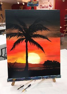 an easel with a painting of a palm tree in front of a sunset background