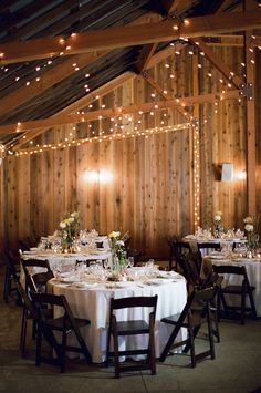 the tables are set with white tablecloths and lights strung from the wooden ceiling